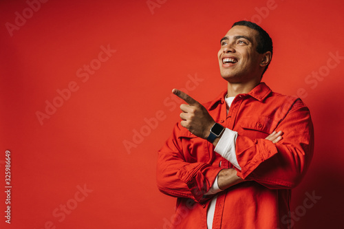 Positive happy young arabian man talking and smiling. Pointing forward. One hand is crossed. Wear colorful clothes. Posing on camera. Isolated over red background. Side view.