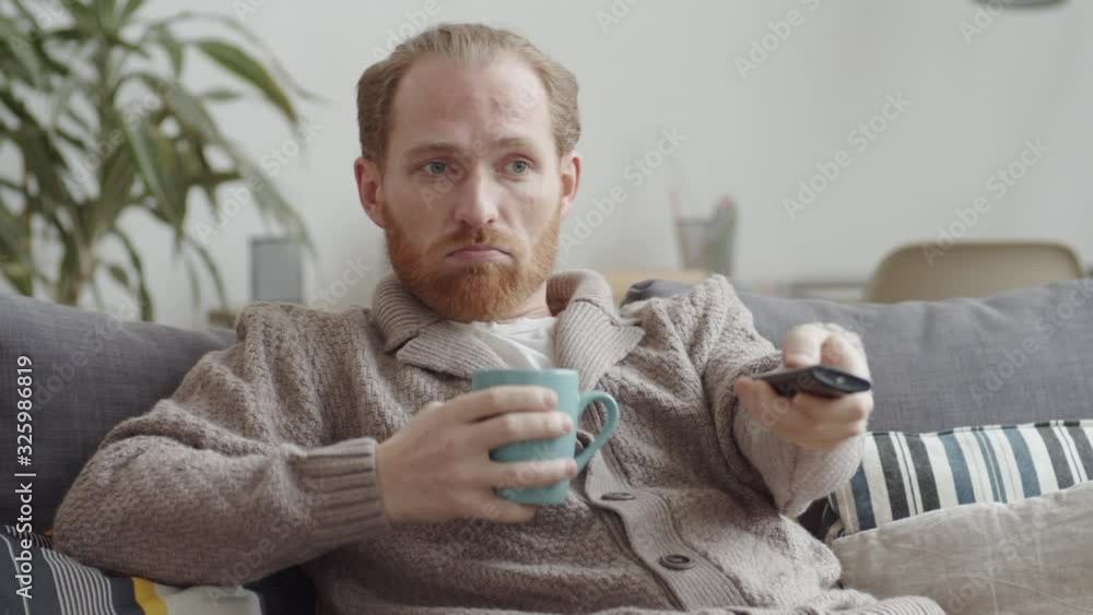 Red-haired Caucasian man lying on couch in front of TV and drinking hot tea Red-haired Caucasian man lying on couch in front of TV and drinking hot tea