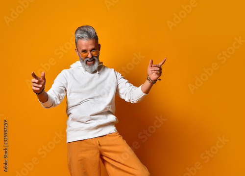 Elderly male in white hoodie, pants and sunglasses, bracelets. Raised hands up, smiling, posing on orange background. Close up
