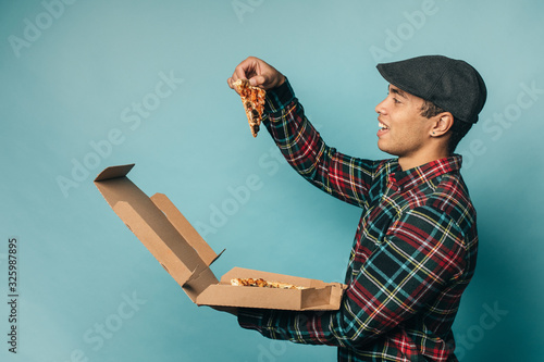 Picture of young man from delievery service stand and hold box with tasty delicious pizza. Take one slice of it and want to eat. Stand alone. Side view. Isolated over blue background. photo