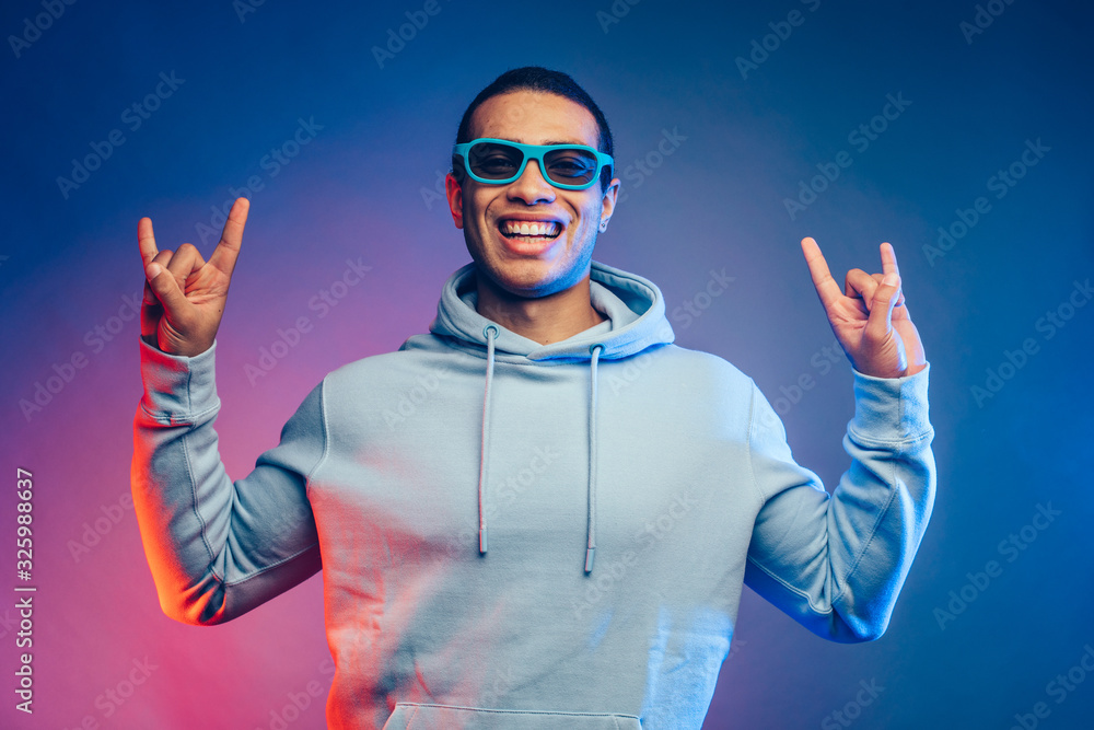 Cool positive and cheerful young egyptian arabian man on picture. Shows rock sign with fingers on both hands. Havin fun alone. Look straight through sunglasses. Isolated over colorful background.