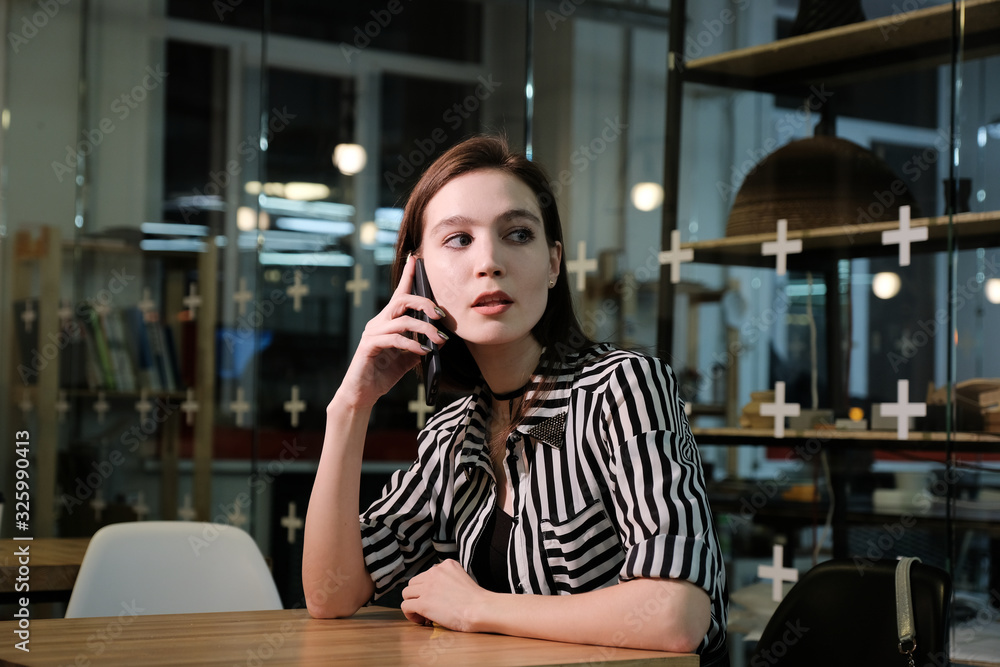 Stylish young beautiful smiling girl in a striped shirt and black dress is sitting at a table in a cafe and talking on the phone. 