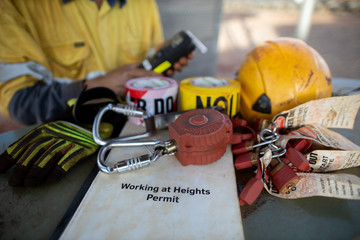 Working at heights permit book placing on the table an inertia reel shock absorbing fall protection device danger caution tags hard hat safety glove and worker holding alcohol tester at the background