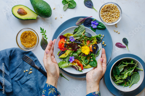 Young woman preparing fresh healthy salad with edible flowers, green, avocado, tomato and pine nuts