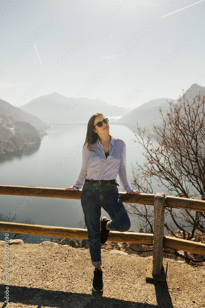 Stylish girl with glasses and blue shirt on top of the hill with lake Lugano in Switzerland at background. She admire the panorama. Girl standing top of monte Bre. Relax day. Stylish girl with glasses and blue shirt on top of the hill with lake Lugano in Switzerland at background. She admire the panorama. Girl standing top of monte Bre. Relax day.