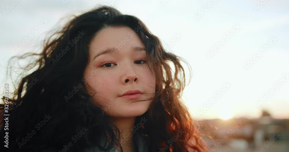 Close up portrait of charming Asian face of beautiful young natural female outdoor, serene teen girl with curly hair looking to camera with sunset skyline light bokeh and city view on background Close up portrait of charming Asian face of beautiful young natural female outdoor, serene teen girl with curly hair looking to camera with sunset skyline light bokeh and city view on background