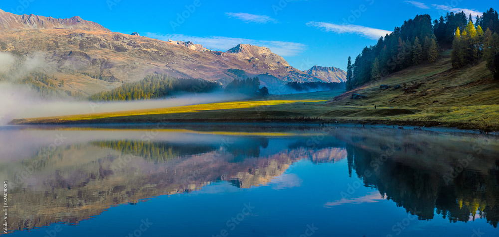 Mountain lake at sunrise in autumn. Landscape with lake, gold sunlight, blue fog over the water, reflection, trees with colorful leaves
