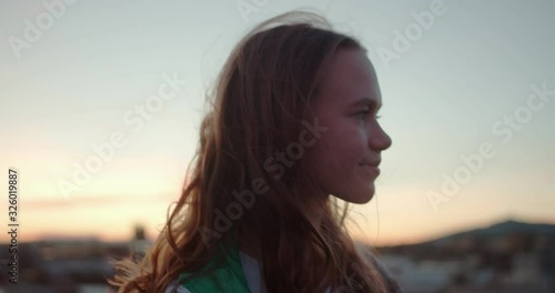 Close up portrait of happy smiling Caucasian young female standing on rooftop outdoor, joyful teen girl enjoying sunset time peaceful candid moment alone city urban view outside windy summer
