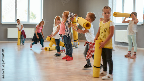 Fototapeta Naklejka Na Ścianę i Meble -  Waiting for the teacher. Group of cute children in casual clothes holding yoga mats and waiting for yoga class in the dance studio