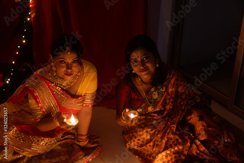 Two beautiful Indian Bengali women in Indian traditional dress lightening Diwali diya/lamps sitting on the floor indoor in darkness on Diwali evening. Indian lifestyle and Diwali celebration