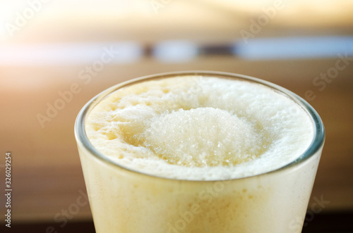 Tall transparent glass beaker, with foam and sugar latte inside, on a wooden table in a cafe in the sunshine