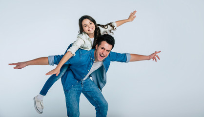 Goofing around. Young handsome father in denim outfit is riding his happy daughter on his back, while they are goofing around together.