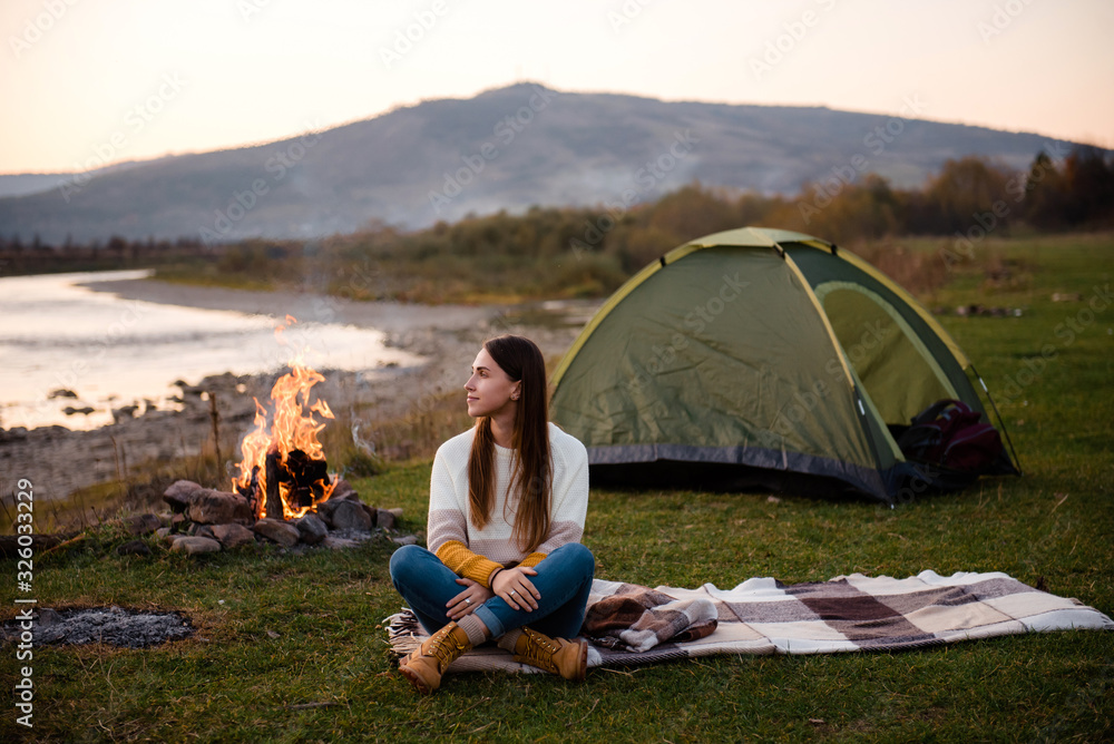 Front view of young brunette in the afternoon sits near a tent and enjoys mountain views. The campfire is lit on the side. Camping and hiking in the mountains