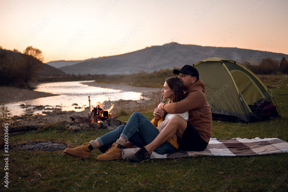 Romantic couple relaxing by the river, enjoying the landscape in the evening. Man and woman sitting on the grass on a blanket and hugging. Near them is a tent and campfire. Mountains in the background