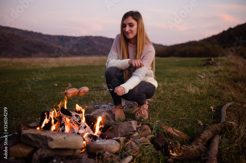 Camping in nature. Beautiful girl on a hike. A woman prepares sausages on a metal skewer on a fire. Green meadow and mountains on the background. Hiking and resting concept