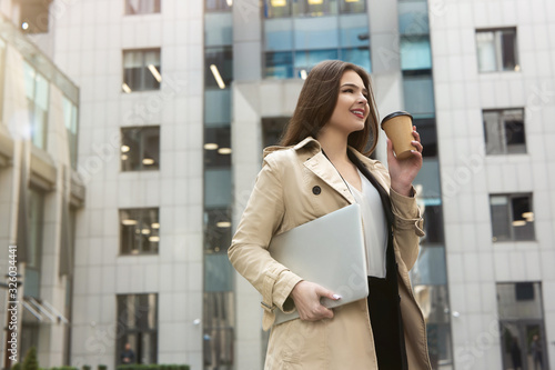 Smiling business lady holding laptop under arm walking down the street near office center with cup of coffee looking pleased, eating on the go concept
