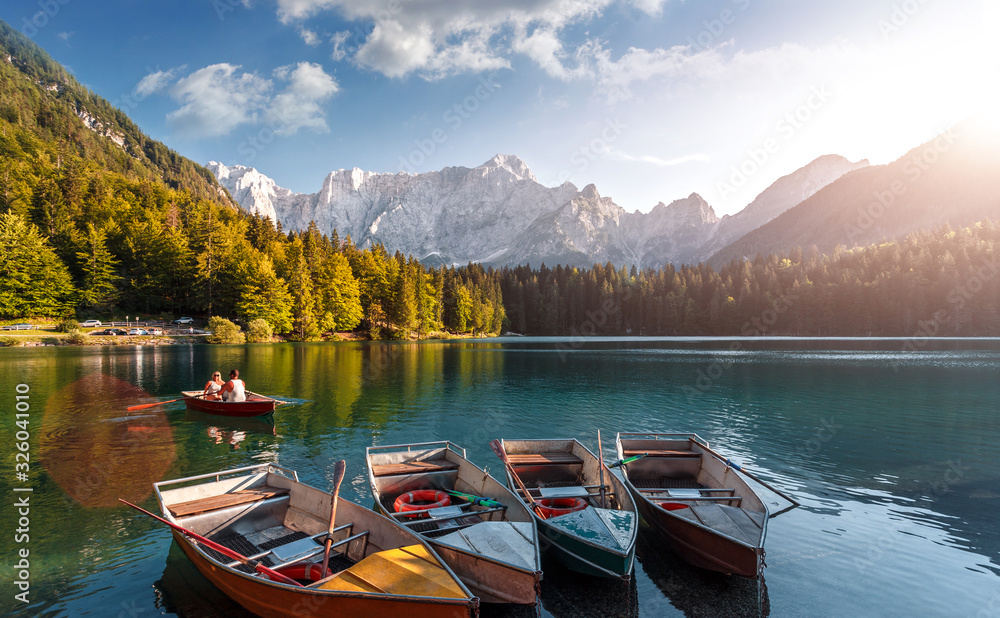 Amazing nature landscape with fairy tale alpine mountain lake Fusine. Beautiful sunny day with blue sky and clouds in summer. Fantastic sunset over the Splendid lake with colorful boats in autumn.