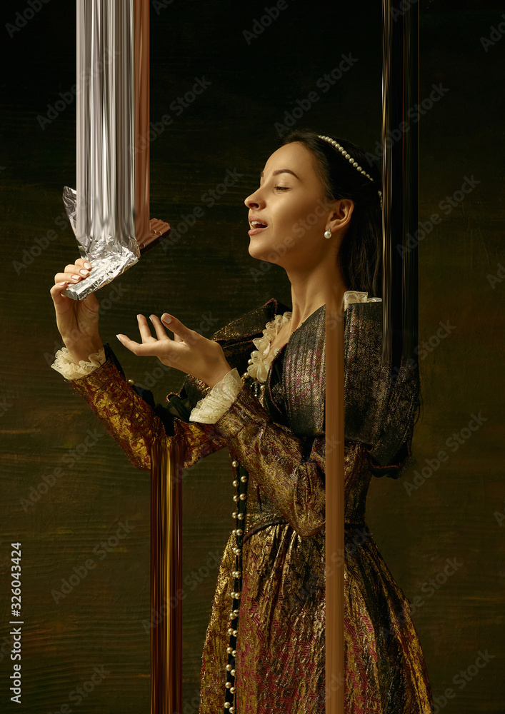 Fototapeta premium Medieval young woman in brown vintage clothing as a duchess on dark studio background. Trying chocolate, sweets. Concept of comparison of eras. Stylish creative design, art vision, new look of artwork