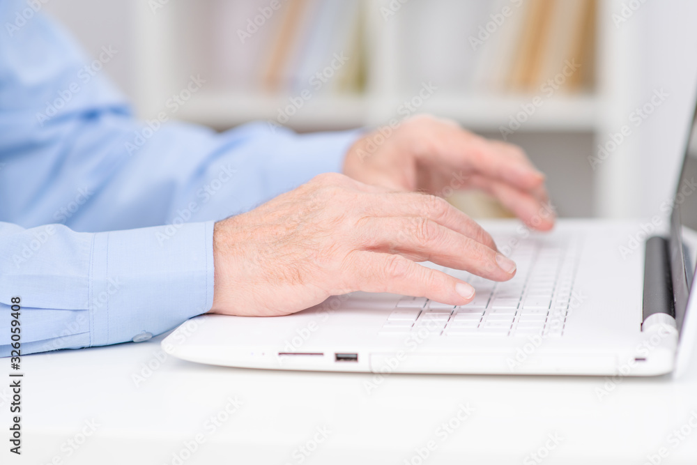 Hands of a senior lying on the keyboard of a white laptop on the background of shelving with books Hands of a senior lying on the keyboard of a white laptop on the background of shelving with books