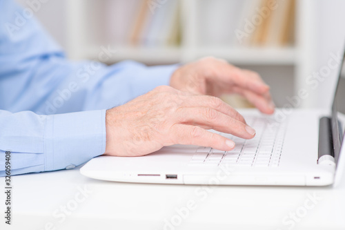 Hands of a senior lying on the keyboard of a white laptop on the background of shelving with books