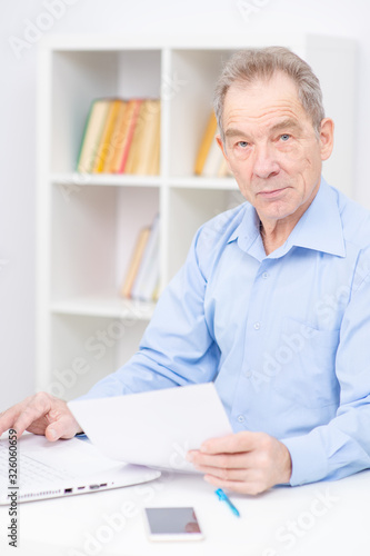 Senior sitting near a laptop, holding papers in his hand and looking at camera.