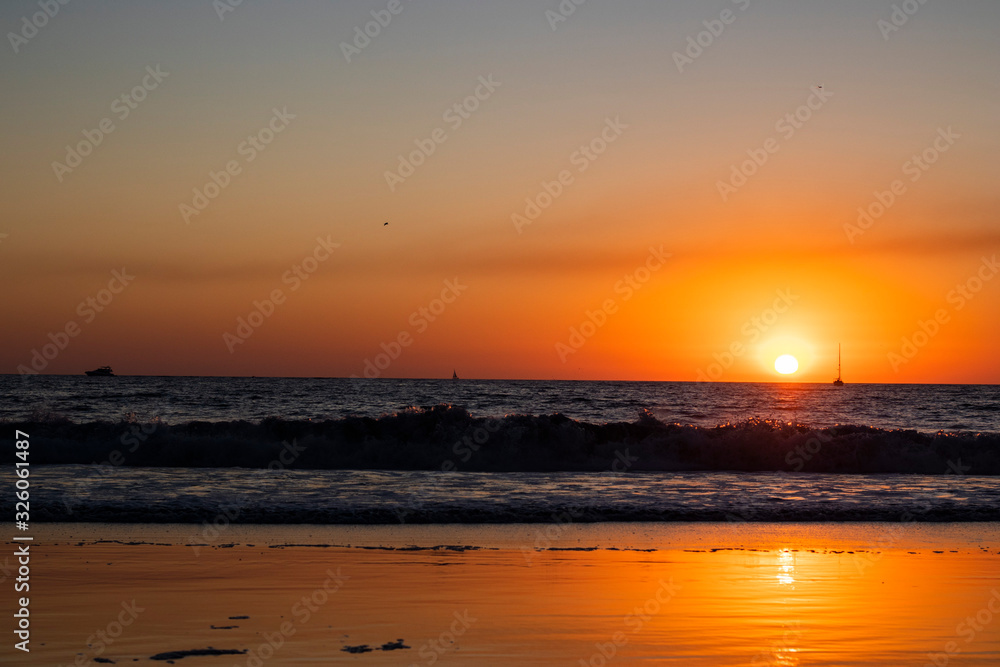 Horizontal image of the sunset over the Pacific Ocean in California with sailboats in the background, reflection of the sun on the sand and close up on the waves in the water