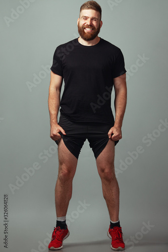 Sportswear concept. Full length portrait of sweet smiling charismatic muscular 30-year-old young man with red hair posing over light gray background. Perfect haircut. Studio shot