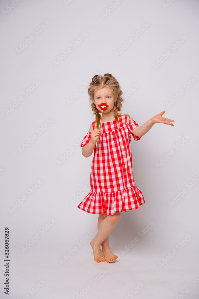 Funny smiling baby girl in a summer dress on a white background full-length . Baby girl with a lip accessory on a stick. Funny smiling baby girl in a summer dress on a white background full-length . Baby girl with a lip accessory on a stick.