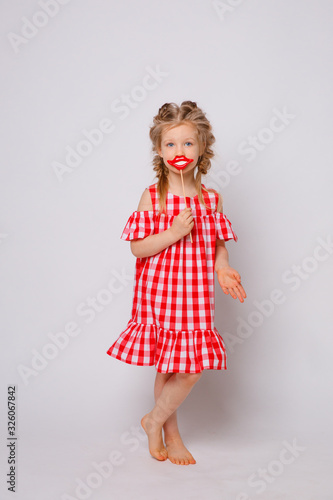 Funny smiling baby girl in a summer dress on a white background full-length . Baby girl with a lip accessory on a stick.