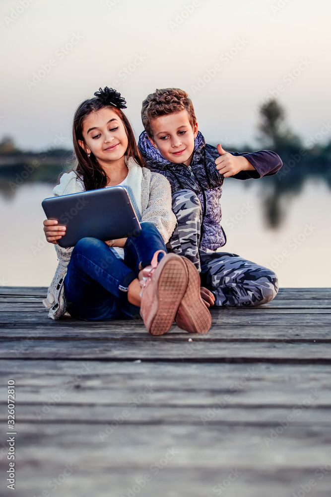 Two happy little friends, boy and girl having fun playing on digital tablet outdoor