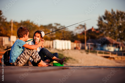 Two young cute little friends, boy and girl talking, eating sandwiches and fishing on a lake in a sunny summer day