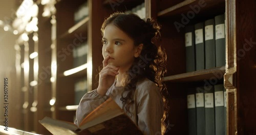 Little beautiful Caucasian girl reading book of fairytales in dark library and dreaming. Cute kid smiling happily while flipping pages and watching pictures of tales in bibliotheca. photo