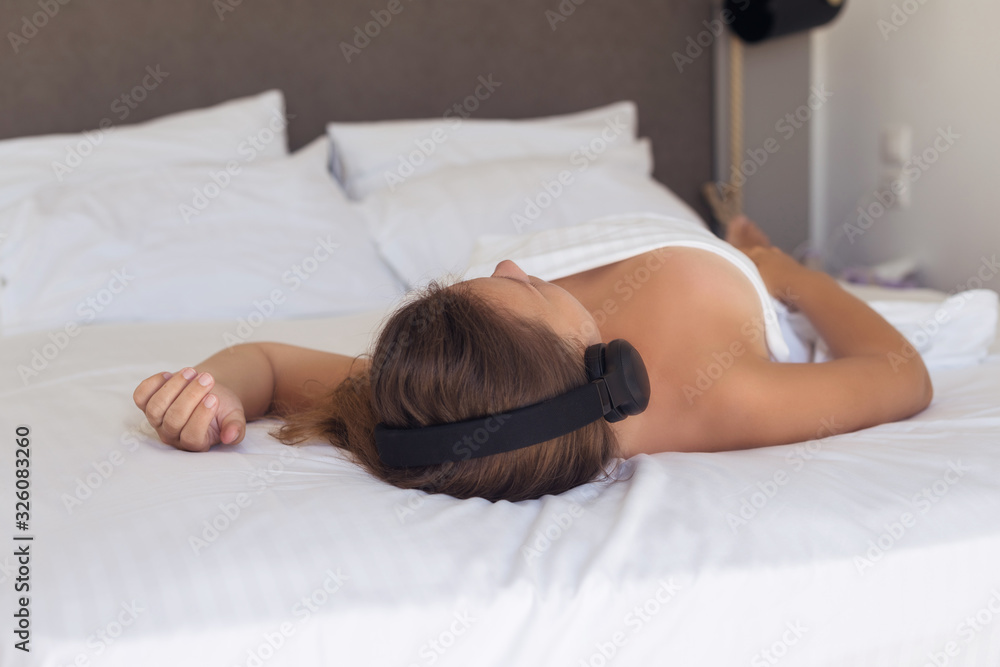 Obraz premium Beautiful naked woman in headphones listening music lying on a bed in the bedroom and covered sheet.