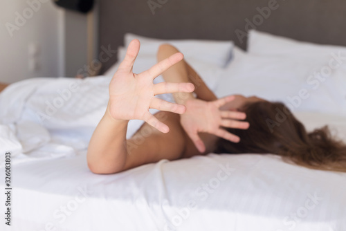 Young woman hides her face from the camera turning her head away lying on a bed in the bedroom and covered sheet