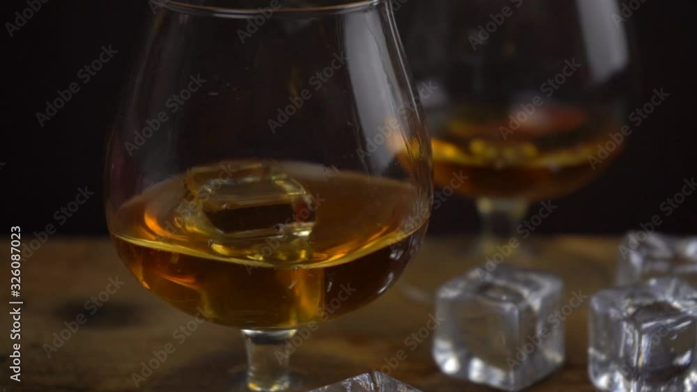 Ice cube falling into cognac \ whiskey glass, golden color strong spirit alcohol in cognac glasses on dark black background, slow motion footage Ice cube falling into cognac \ whiskey glass, golden color strong spirit alcohol in cognac glasses on dark black background, slow motion footage