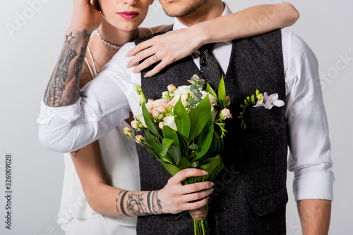 cropped view of tattooed bride embracing bridegroom isolated on grey