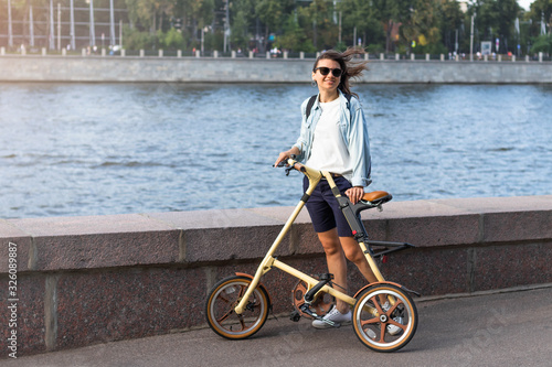 Beautiful happy white woman in sunglasses stands on the embankment of a wide river. In the hands of a folding comfortable yellow bike. Summer or spring life style.