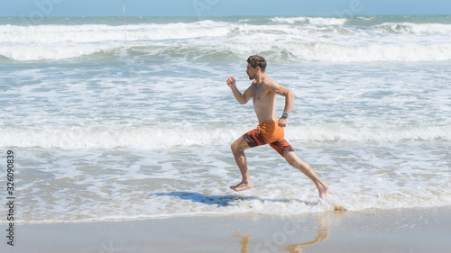 Handsome man with athletic body running around the beach line near the sea.Fit runner on the beach with sea waves background. Beach activities concept.