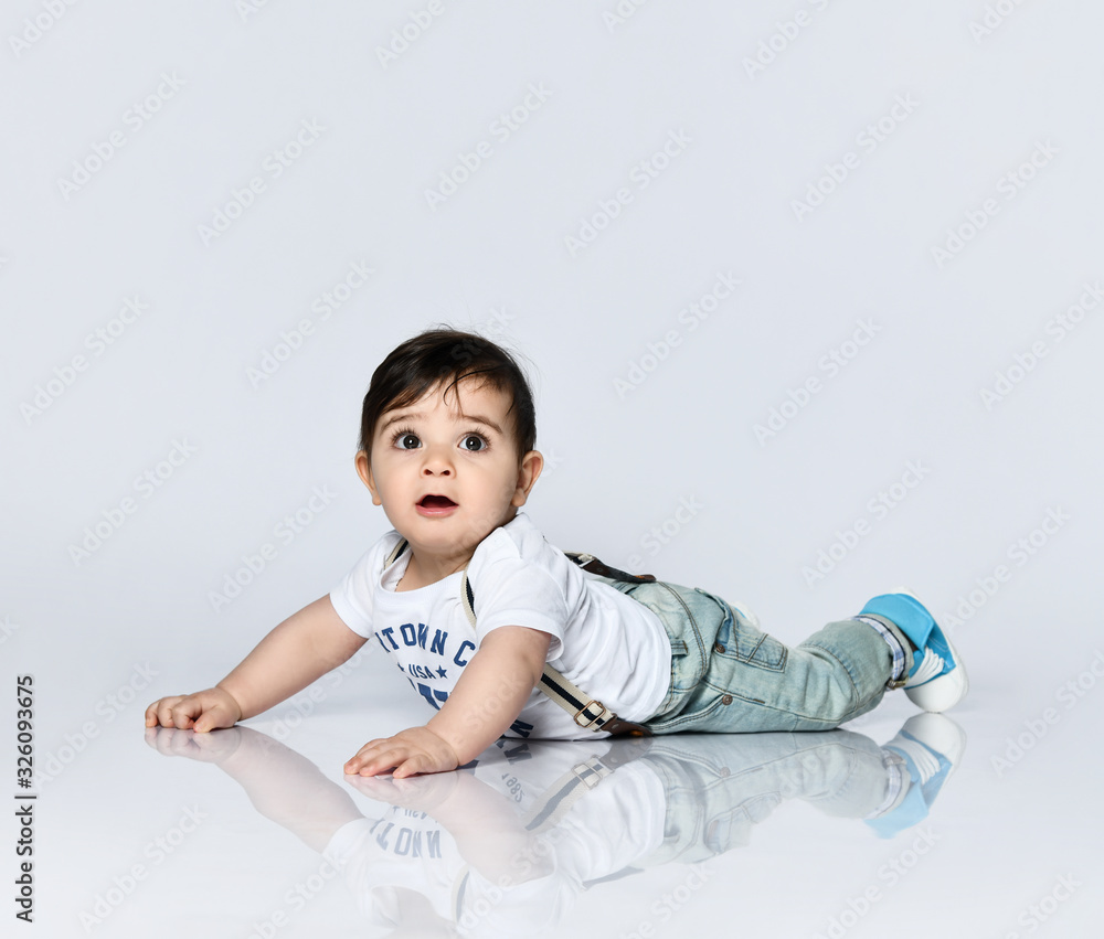 Little toddler in t-shirt with inscriptions, jeans with suspenders. He is laying on floor isolated on white background. Close up Little toddler in t-shirt with inscriptions, jeans with suspenders. He is laying on floor isolated on white background. Close up