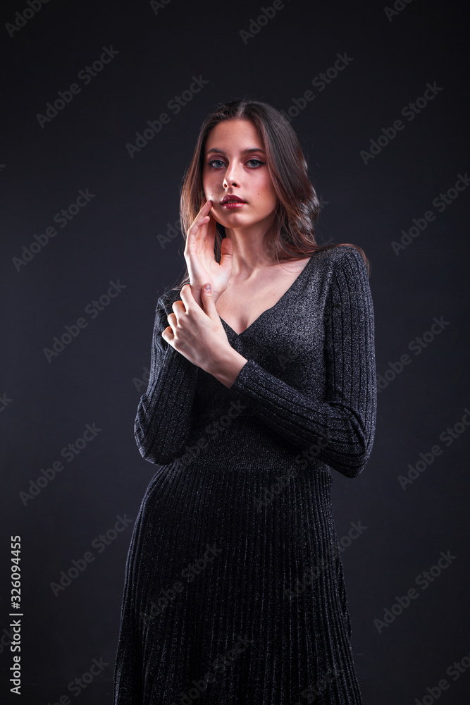 Fototapeta premium Portrait of a young brunette girl in black tight dress on a black wall backgroundll
