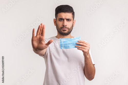 Portrait of man warning of virus danger, gesturing stop and holding protective hygienic mask to prevent coronavirus infection, respiratory diseases such as flu, 2019-nCoV. indoor studio shot, isolated © khosrork