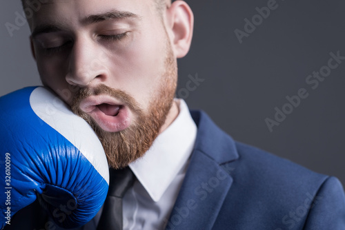 Fotografie close-up portrait of a young bearded guy in a business suit, businessman, side view the hand of an opponent in a boxing glove strikes