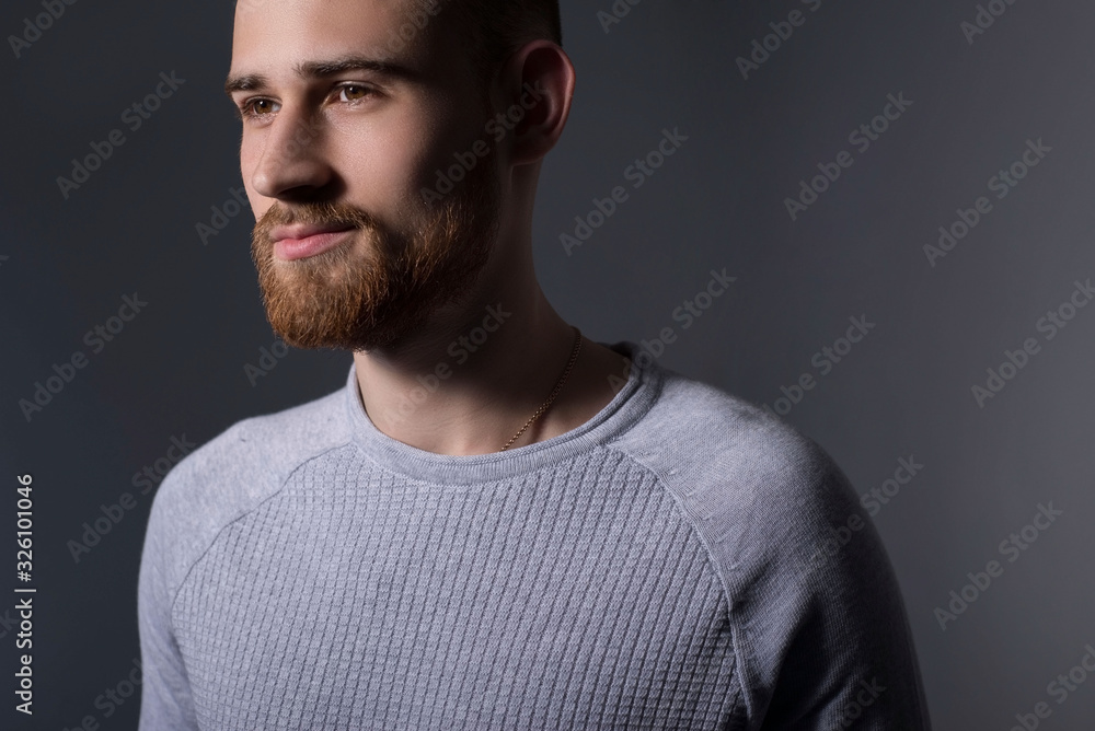 closeup, dramatic portrait of a handsome bearded guy of twenty-five years old, in a gray sweater, on a gray background. Casual style. Sweater, mens jacket. Smiling bearded young guy.
