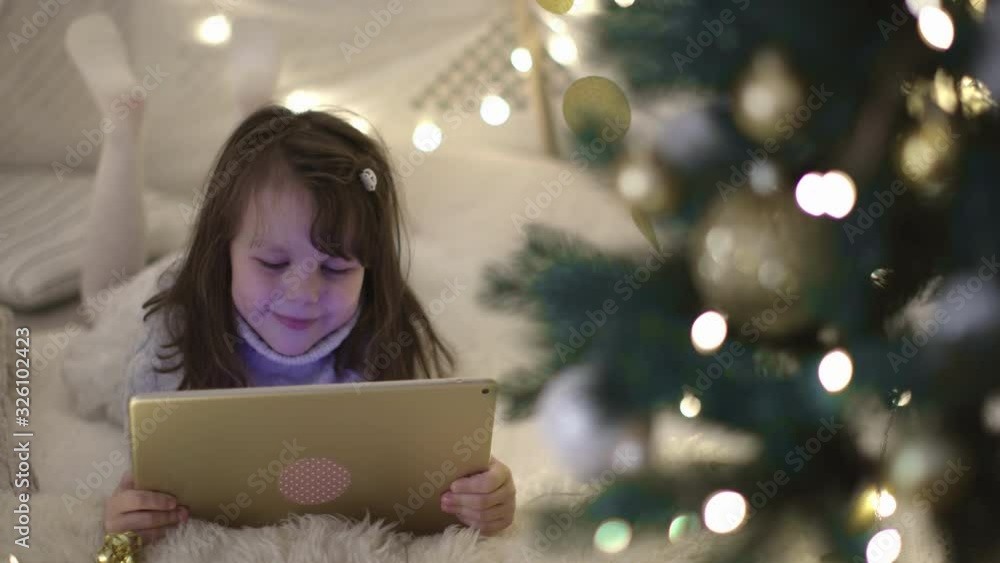 A Little Girl in a Wool Dress Lies on the Carpet and Plays with a Tablet Computer in Christmas Decoration. New Year Family Holiday. A Little Girl in a Wool Dress Lies on the Carpet and Plays with a Tablet Computer in Christmas Decoration. New Year Family Holiday.