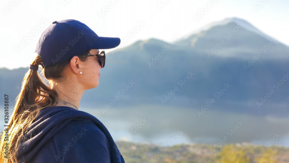 A close up on woman, enjoying the volcanic landscape of Bali, Indonesia, seen from the top of Mount Batur. Woman is wearing a cap and dark sunglasses. Enjoying the moment. Mount Agung in the back A close up on woman, enjoying the volcanic landscape of Bali, Indonesia, seen from the top of Mount Batur. Woman is wearing a cap and dark sunglasses. Enjoying the moment. Mount Agung in the back