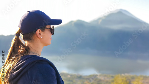 A close up on woman, enjoying the volcanic landscape of Bali, Indonesia, seen from the top of Mount Batur. Woman is wearing a cap and dark sunglasses. Enjoying the moment. Mount Agung in the back © Chris