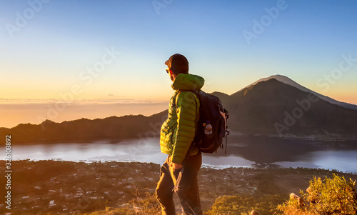 A man in a jacket standing on top of Mount Batur, Bali, Indonesia and waiting for the sunrise. There is Mount Rinjani in the back (Lombok) and Volcano Agung on the side. Fog in the valley. Mysterious