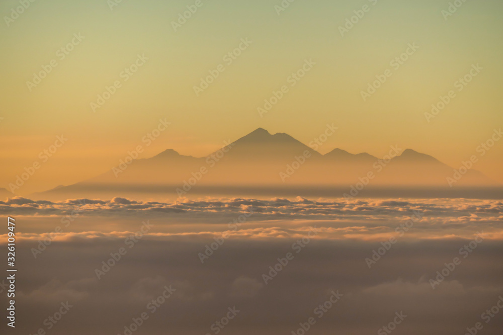 A distant view on Mount Rinjani on Lombok, Indonesia, captured from Mount Batur on Bali, during the morning golden hour. There is a slight fog around the lower parts. Mysterious and magical sunrise.