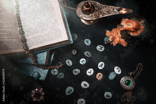 Astrology and esotericism. On a black background lie fortune-telling runes, a book, precious amulets, a copper lamp and a candle. Top view. Dust and light