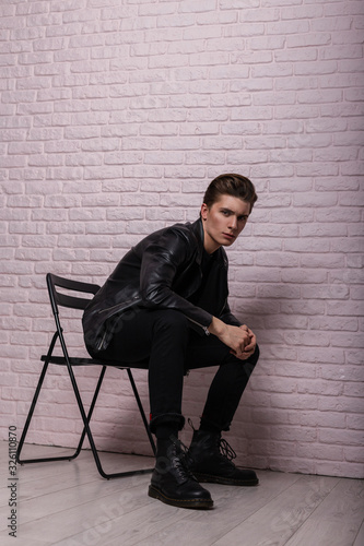 Young handsome man hipster with trendy hairstyle in fashionable black stylish youth clothes in cool leather boots is sist on a vintage chair near a pink brick wall in the studio. American trendy guy. photo